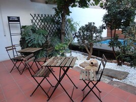 An outdoor patio with wooden tables and chairs, surrounded by plants and a pool.