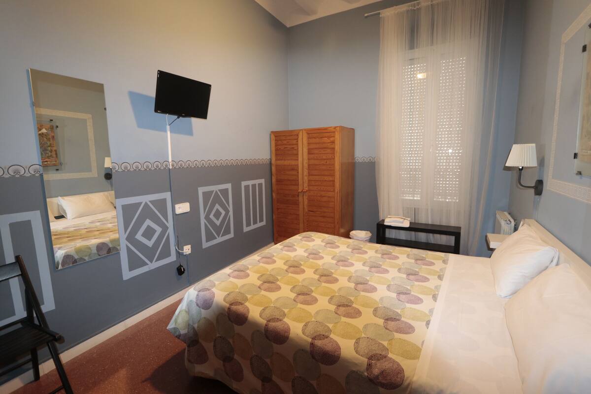 A bedroom with a bed, a wardrobe, a desk, a chair, a TV, and a window with curtains.