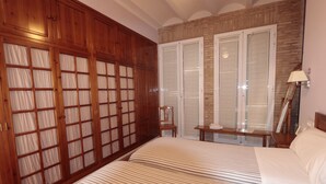 Un dormitorio con un amplio armario de madera, una cama con ropa de cama a rayas, una mesita pequeña y una silla.