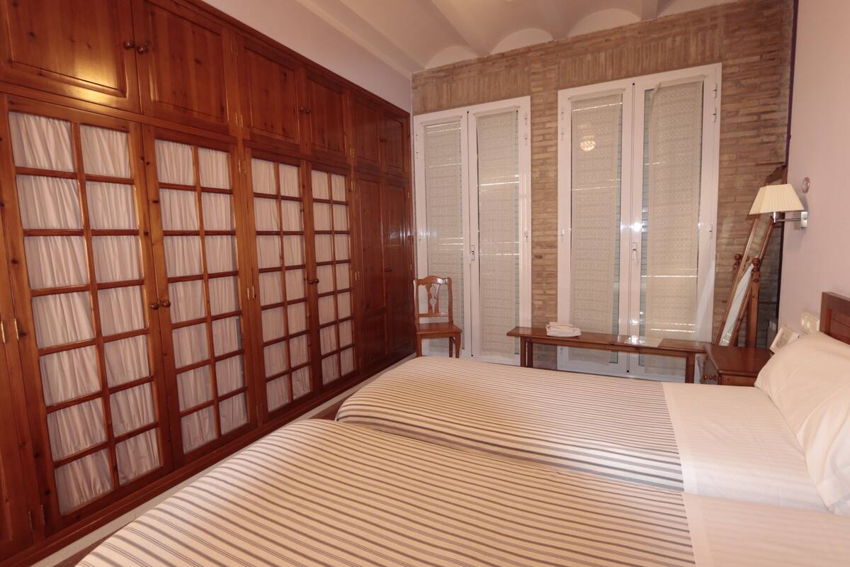 A bedroom with a large wooden wardrobe, a bed with striped bedding, a small table, and a chair.
