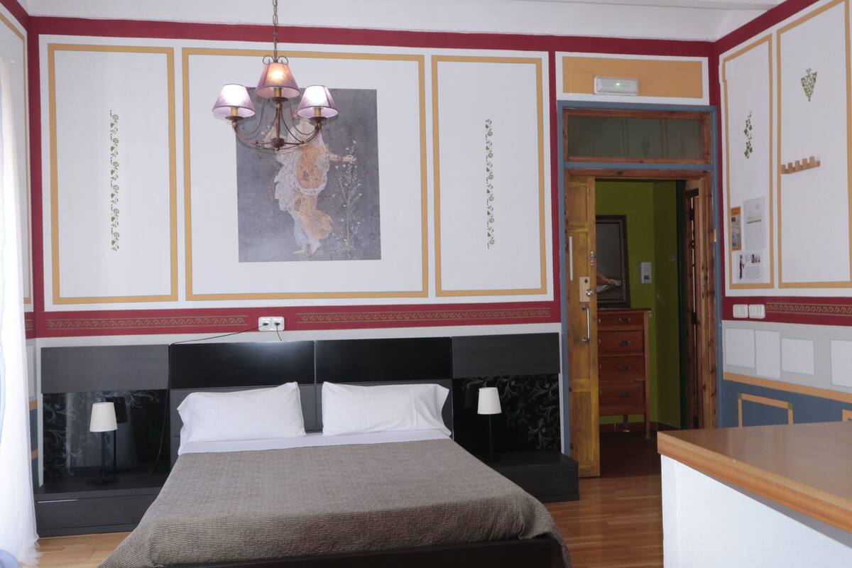 A hotel room with a large bed, a chandelier, a painting on the wall, and a wooden desk.
