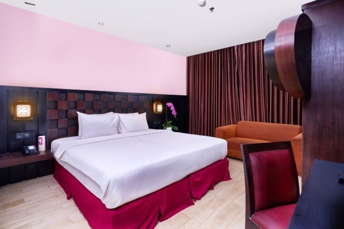 Une chambre d’hôtel moderne dotée d’un grand lit, d’une tête de lit en bois, d’un mur d’accent rouge et d’un coin salon avec un fauteuil et un canapé rouges.
