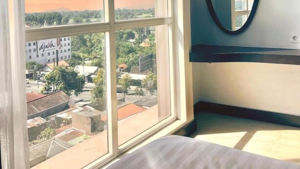 A bedroom with a large window, a bed, a round mirror, and a view of a cityscape. - Grand Orchid Hotel Yogyakarta (Depok)