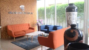 A modern hotel lobby with a brick-patterned wall, a Grand Orchid Hotel sign, a white coffee table, orange and blue sofas, and a large decorative vase. - Grand Orchid Hotel Yogyakarta (Depok)