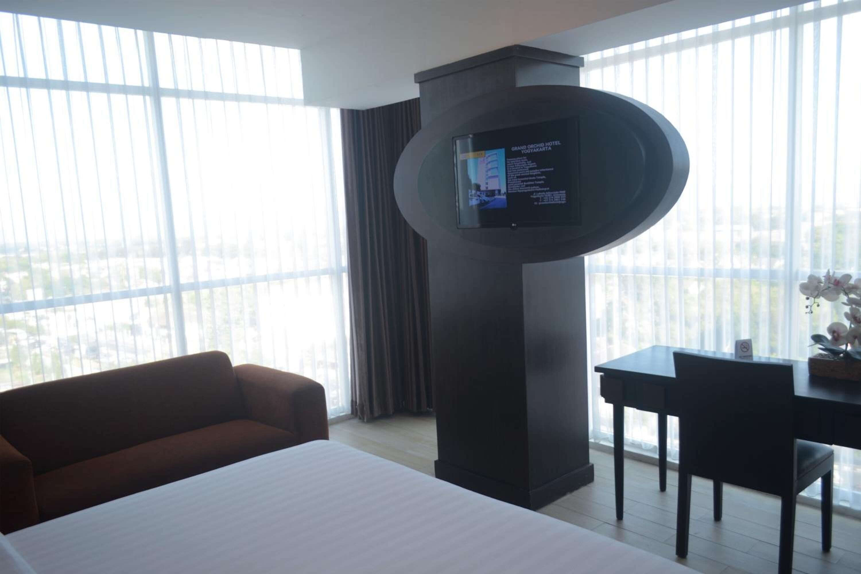 A hotel room with a large bed, a TV mounted on a pillar, a desk with a chair, and a view of the city through large windows.