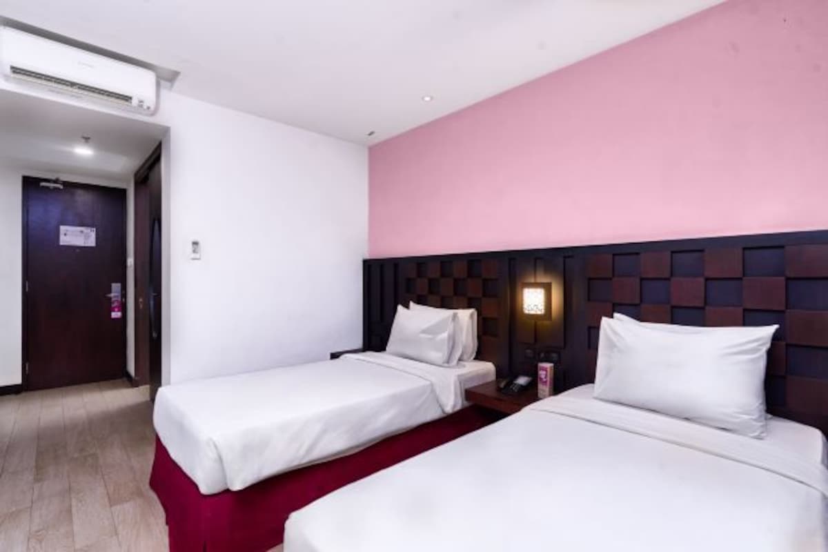 Habitación de hotel con dos camas individuales, alfombra roja, cabecero de madera, mesita de noche con teléfono e iluminación en la pared.