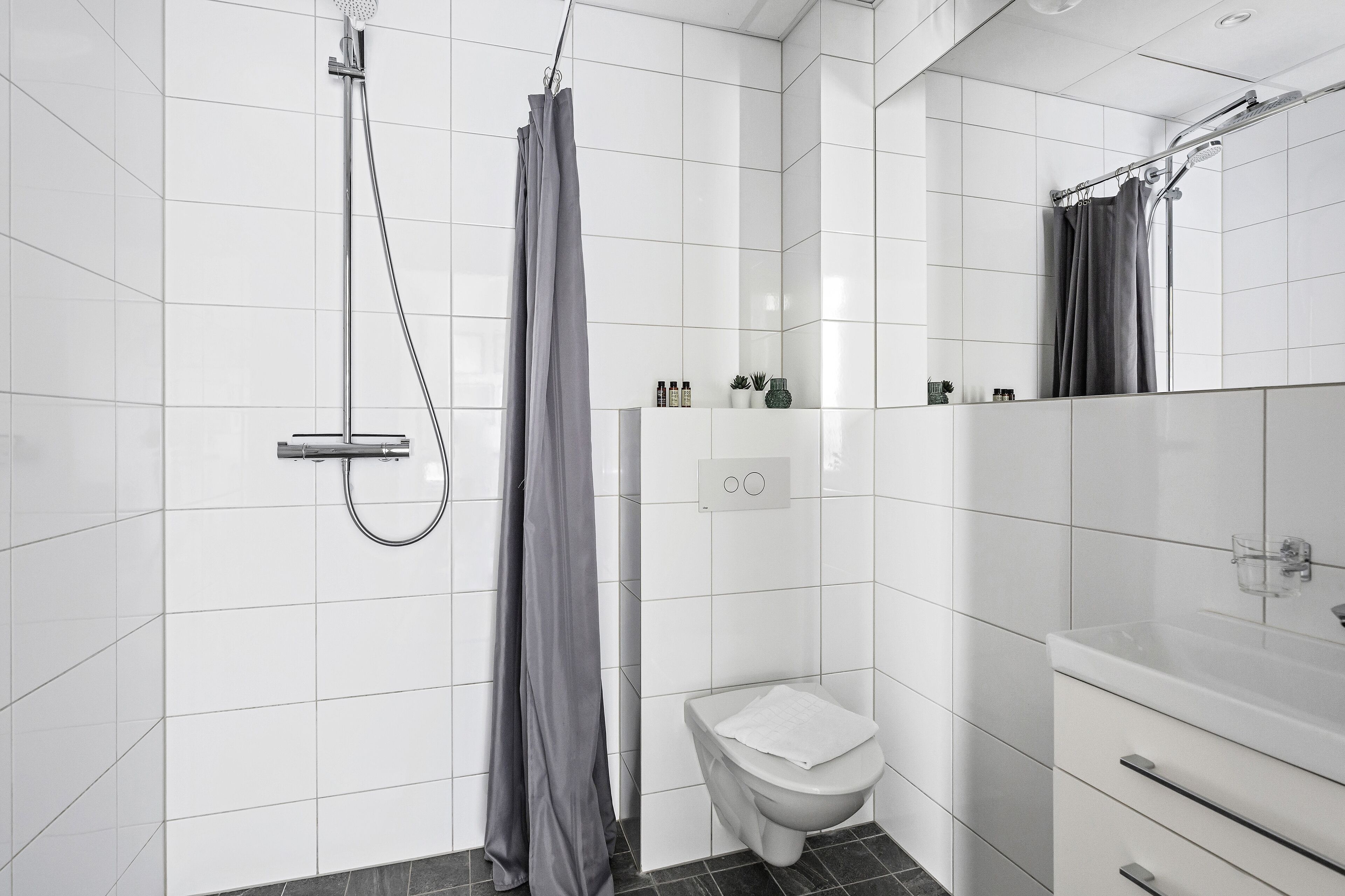 A modern bathroom with white tiles, a shower with a grey curtain, a toilet, and a vanity with a mirror.