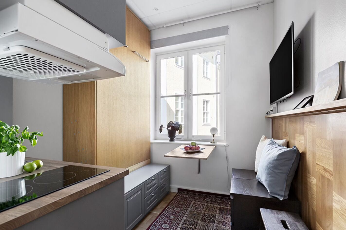 A compact kitchen with a built-in stove, a window with a view, a TV mounted on the wall, and a small table with a bowl of fruit.