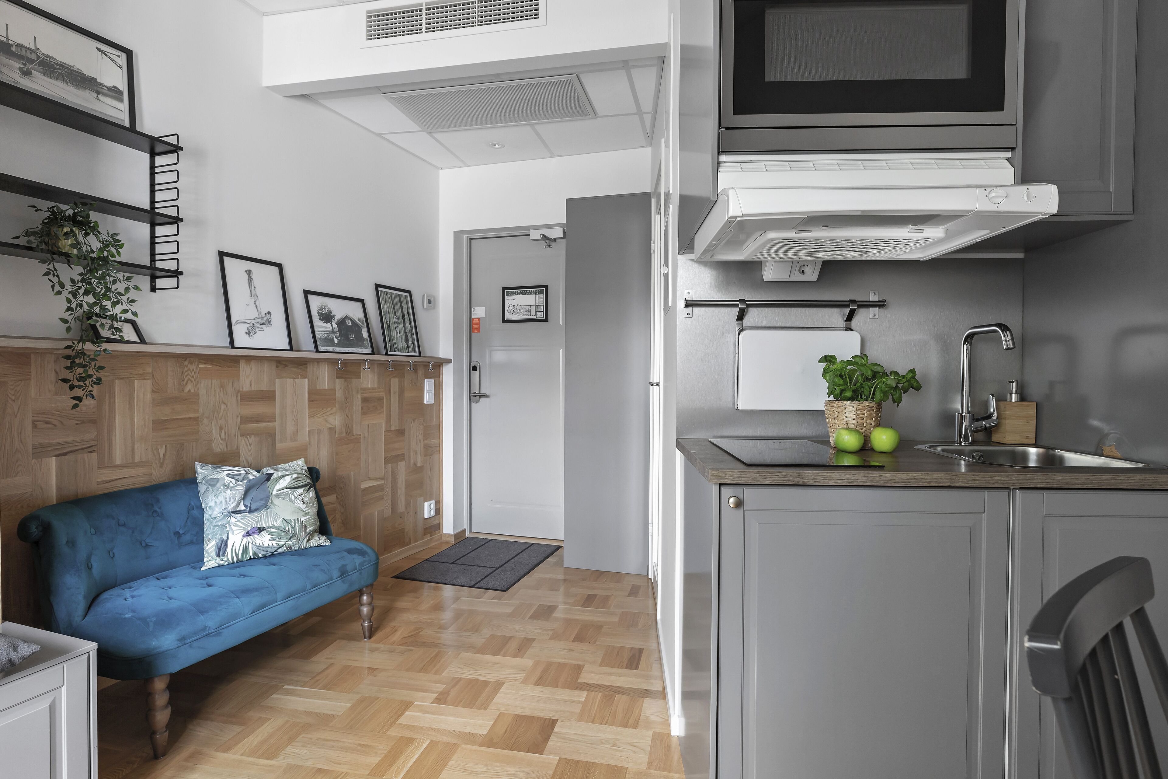 Une cuisine compacte équipée d’un four à micro-ondes, d’un évier et d’un petit plan de travail. Un canapé bleu et un meuble en bois sont visibles dans l’espace de vie.