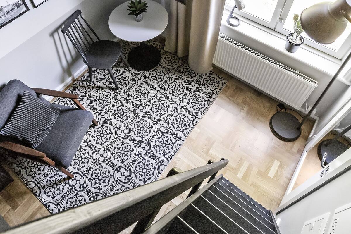 Ein modernes Zimmer mit einem schwarz-weißen Fußboden, einem runden Tisch mit einer Pflanze, einem Sessel, einem Schreibtisch mit einem Laptop, einer Pendelleuchte und einem Fenster mit Vorhängen.