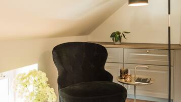Een zwarte beklede stoel, een vloerlamp, een houten bureau en een vaas met bloemen in een kamer met houten vloer en een schuine plafond.