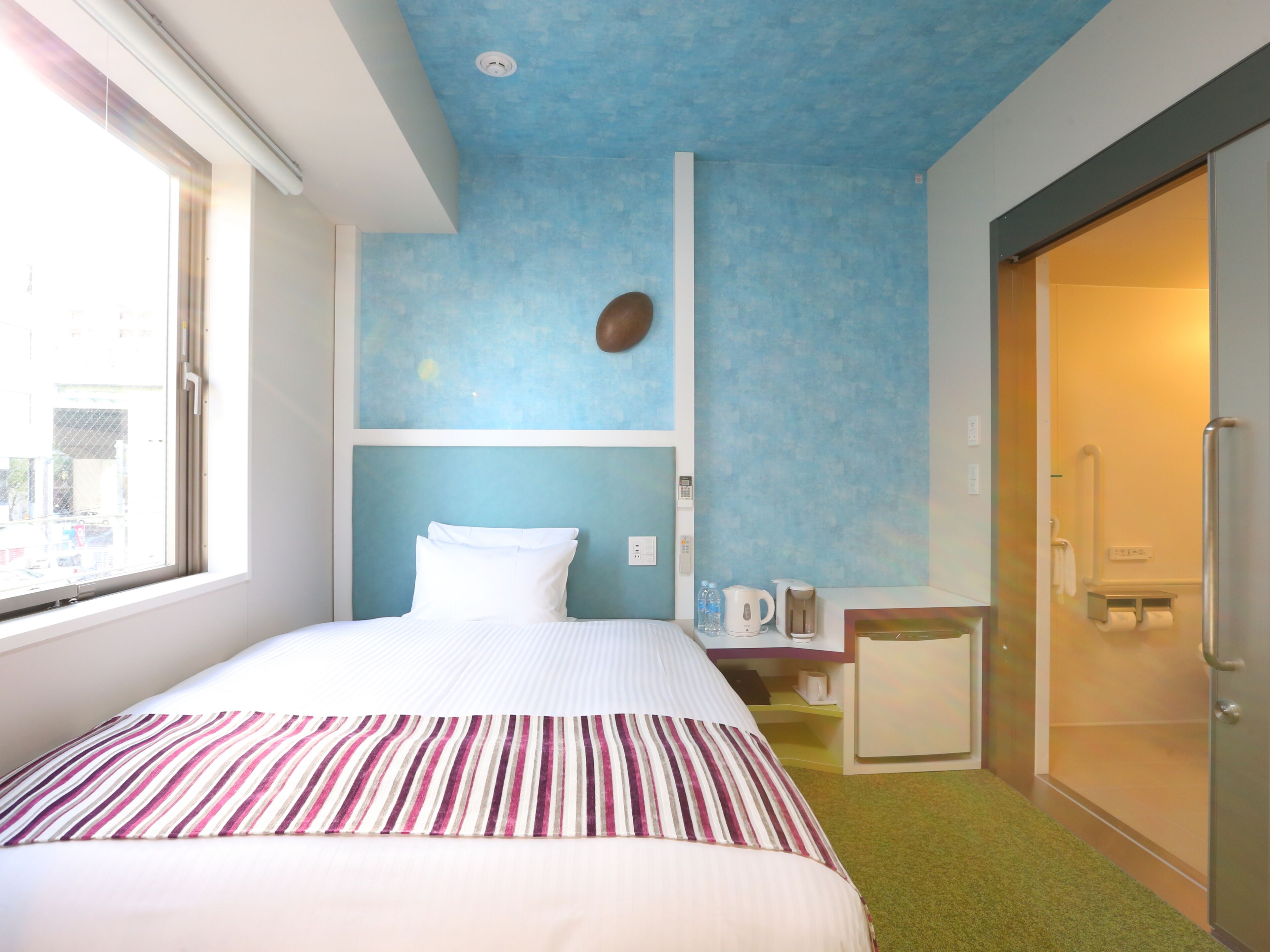 A hotel room with a bed, a small table with a kettle, a shower area, and a view of the outside.