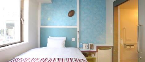 A hotel room with a bed, a small table with a kettle, a shower area, and a view of the outside.