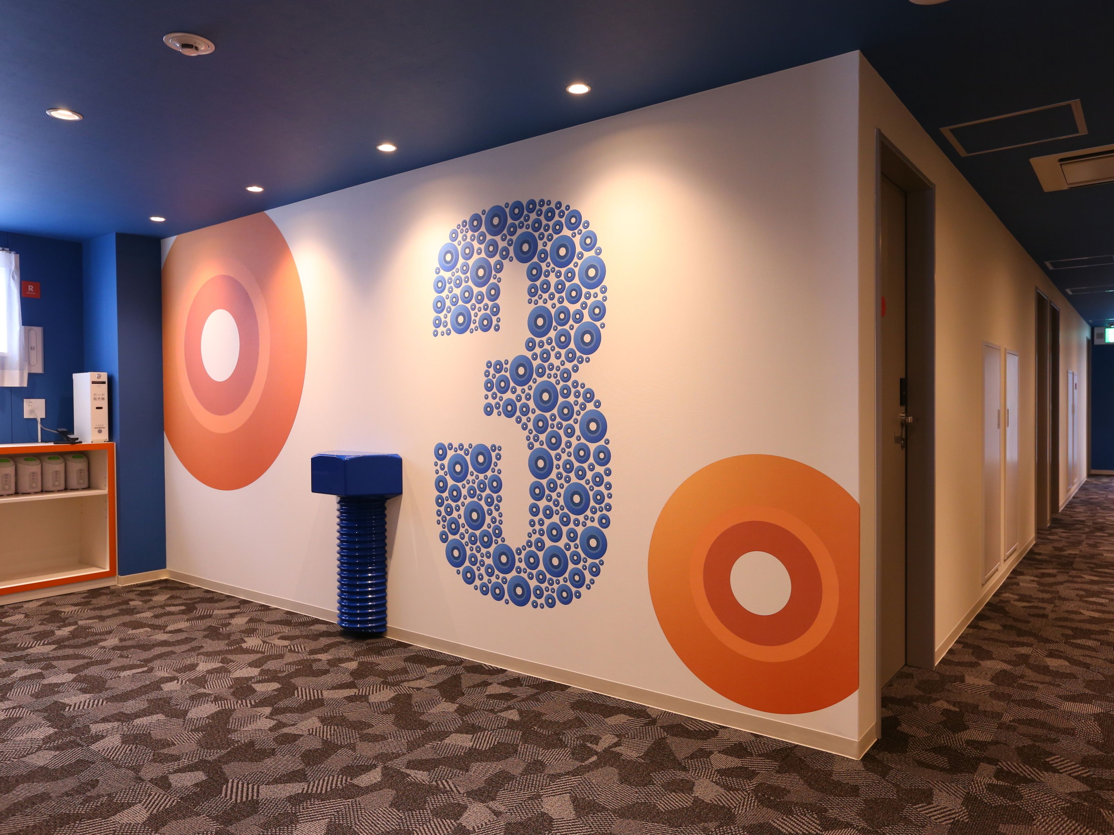 A hallway with a wall featuring a large number '3' design, a blue cylindrical object, and patterned carpet.
