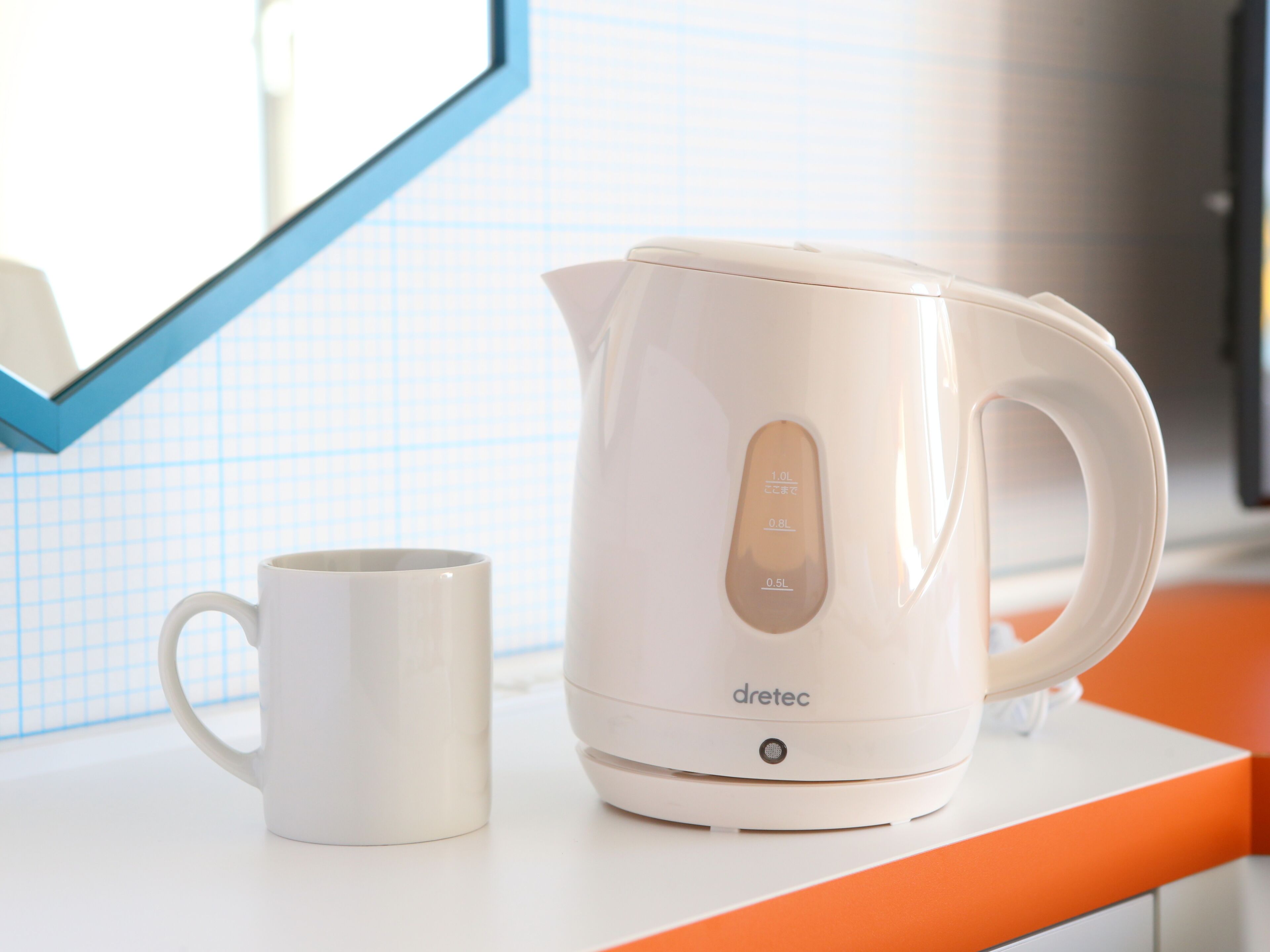 Une bouilloire électrique blanche et une tasse blanche sont posées sur un plan de travail blanc bordé d’une ligne orange.
