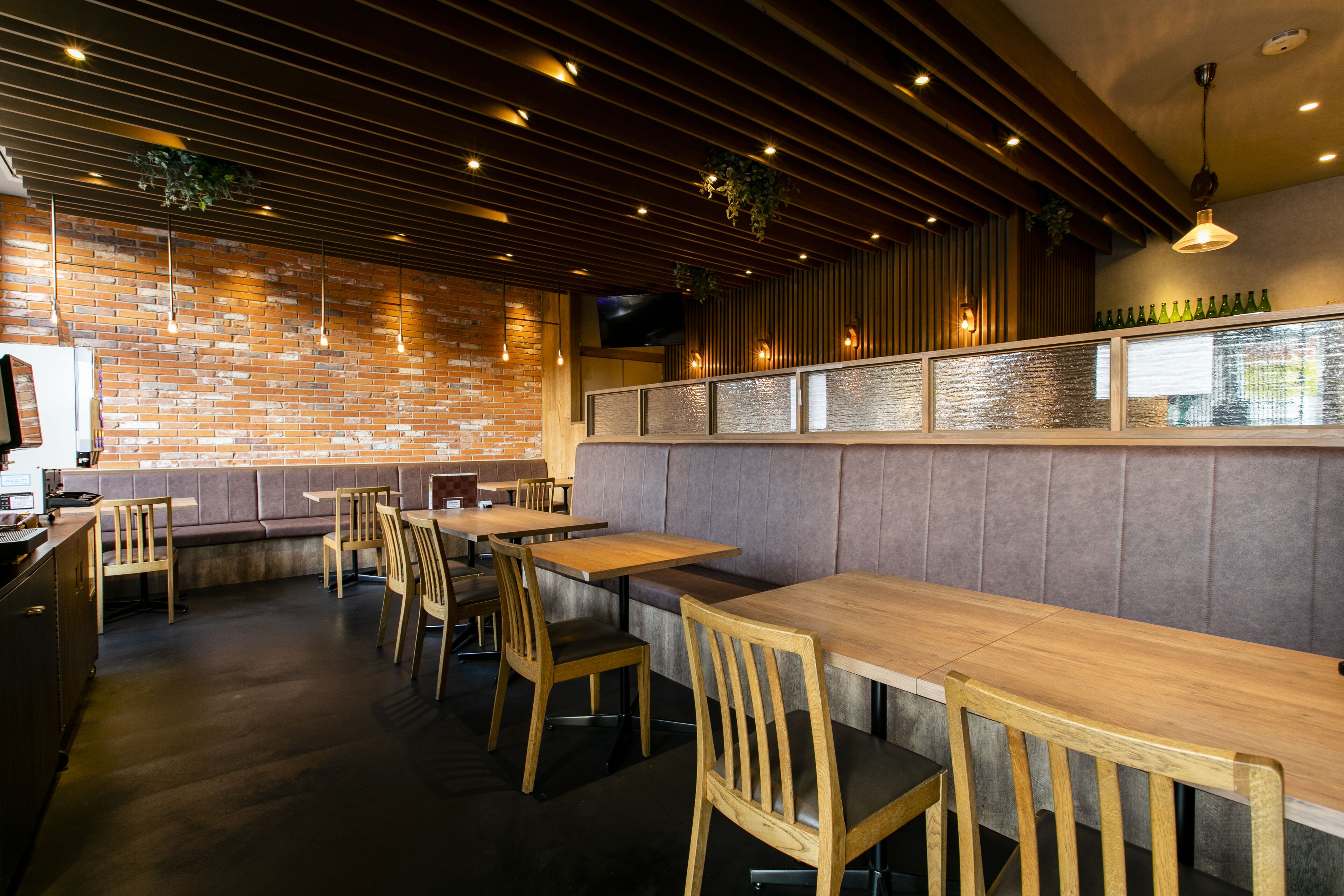 A restaurant interior with brick walls, wooden tables and chairs, and a bar area with a glass partition.