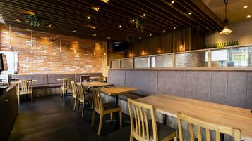 A restaurant interior with brick walls, wooden tables and chairs, and a bar area with a glass partition.