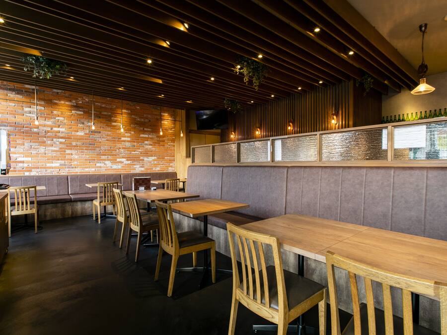 A restaurant interior with brick walls, wooden tables and chairs, and a bar area with a glass partition.