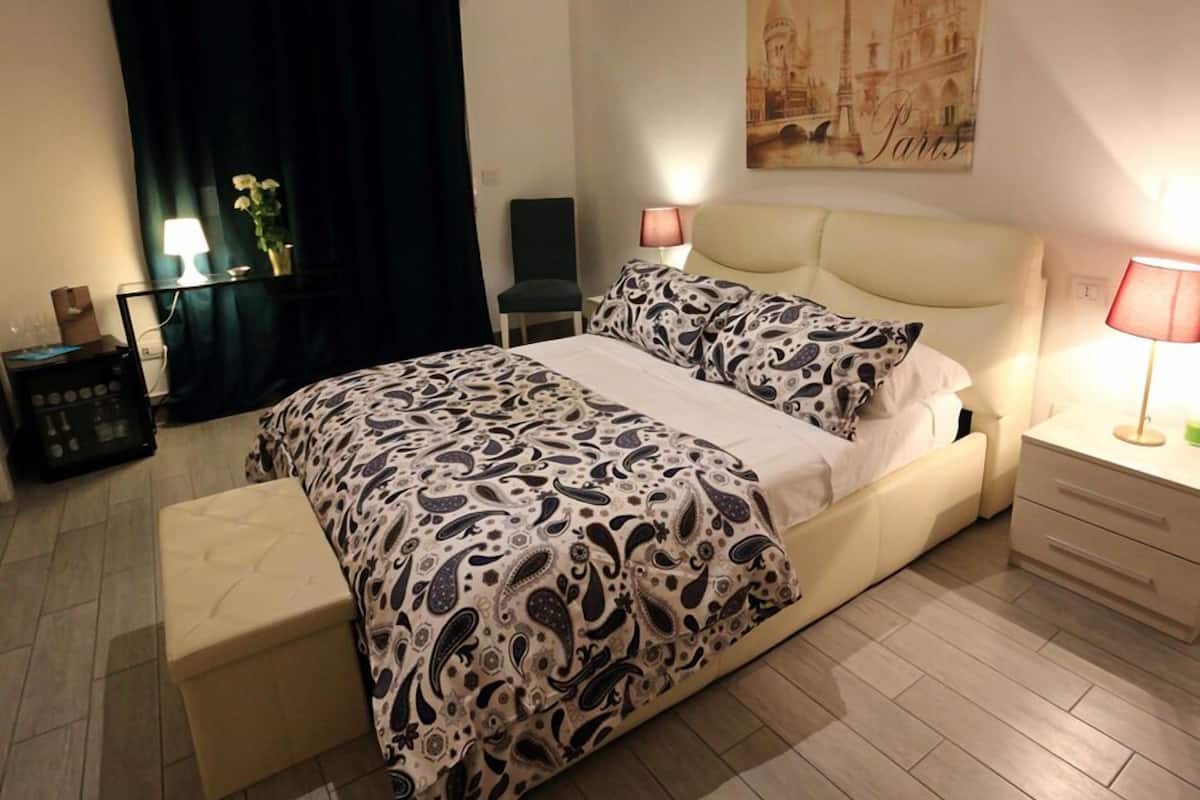 A neatly arranged bedroom with a bed, bedside tables, a chair, a desk, and a wall painting.