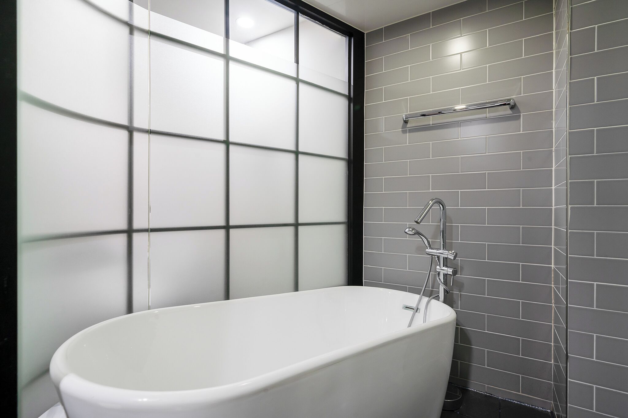 Ein modernes Badezimmer mit freistehender Badewanne, einer Milchglasabtrennung und grauen Fliesenwänden.