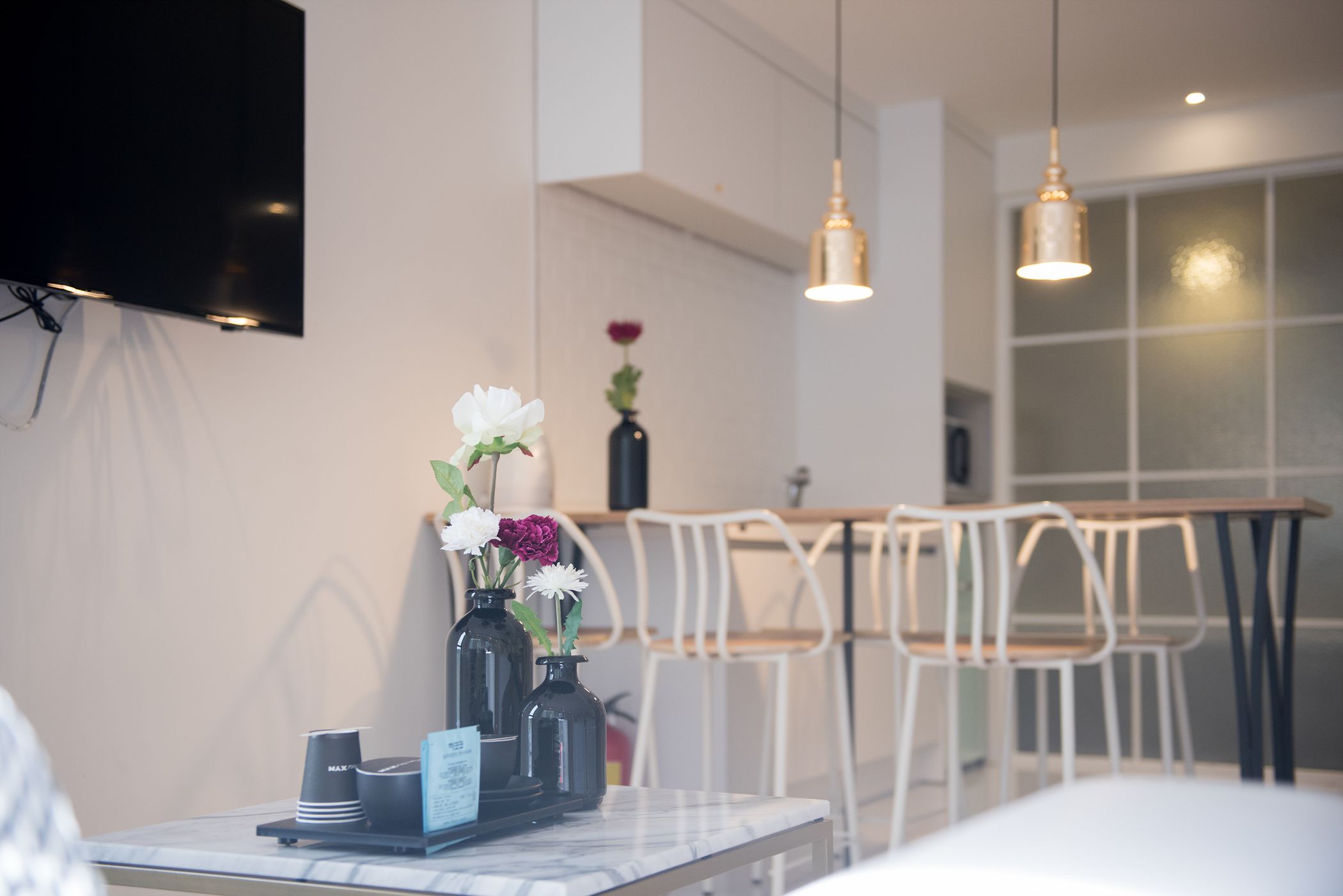 Una habitación de hotel moderna con una televisión de pantalla plana, una mesa de mármol con un florero y una zona de comedor con sillas blancas y una mesa de madera.