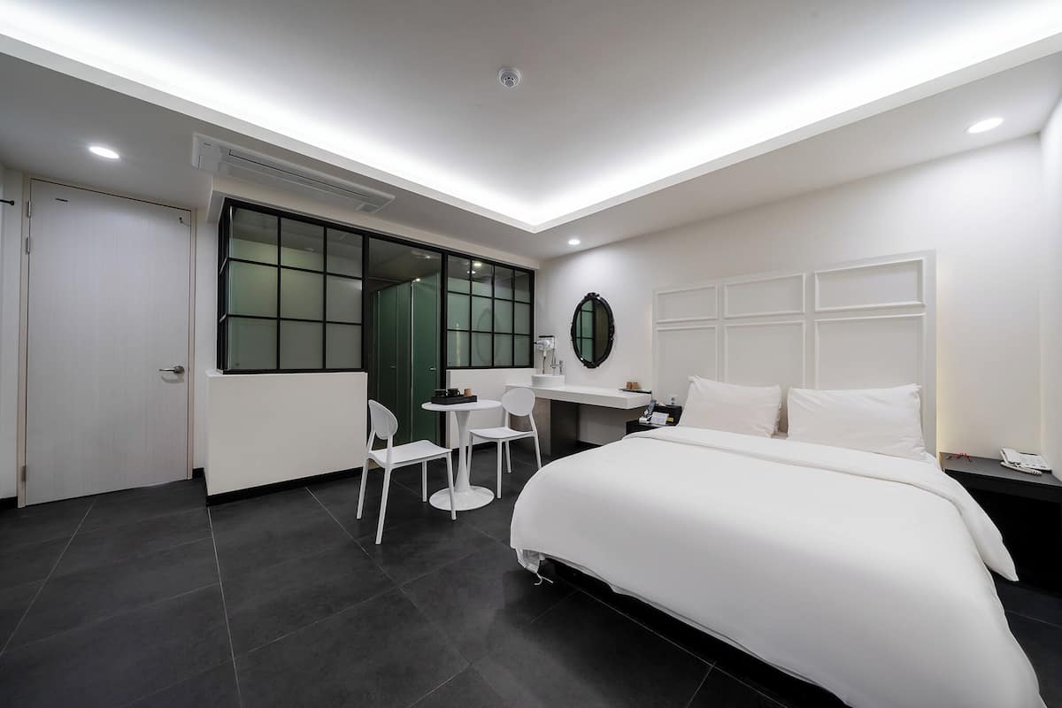 A modern hotel room with a large bed, a desk with a chair, a small table, and a mirror on the wall.