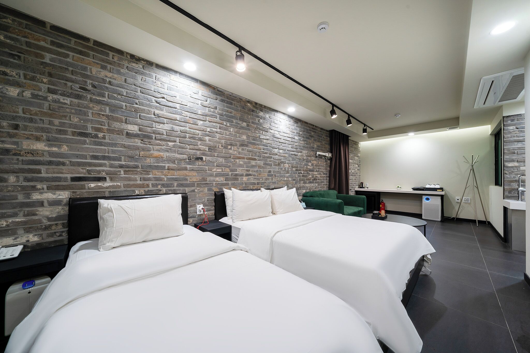 A modern hotel room with two beds, a brick accent wall, a desk with a chair, and a small kitchenette.