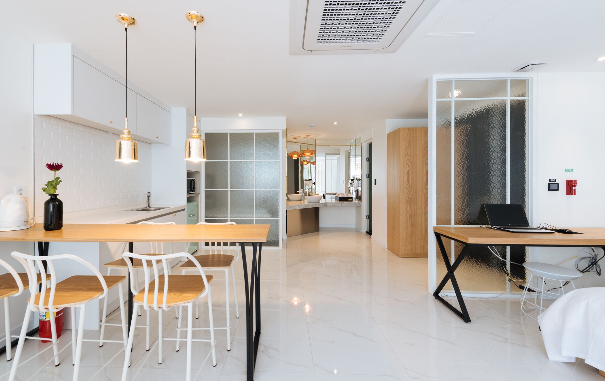 A modern kitchen with white cabinets, a wooden dining table, and white chairs. There is a laptop on a desk with a chair, and a vase with a flower on the counter.