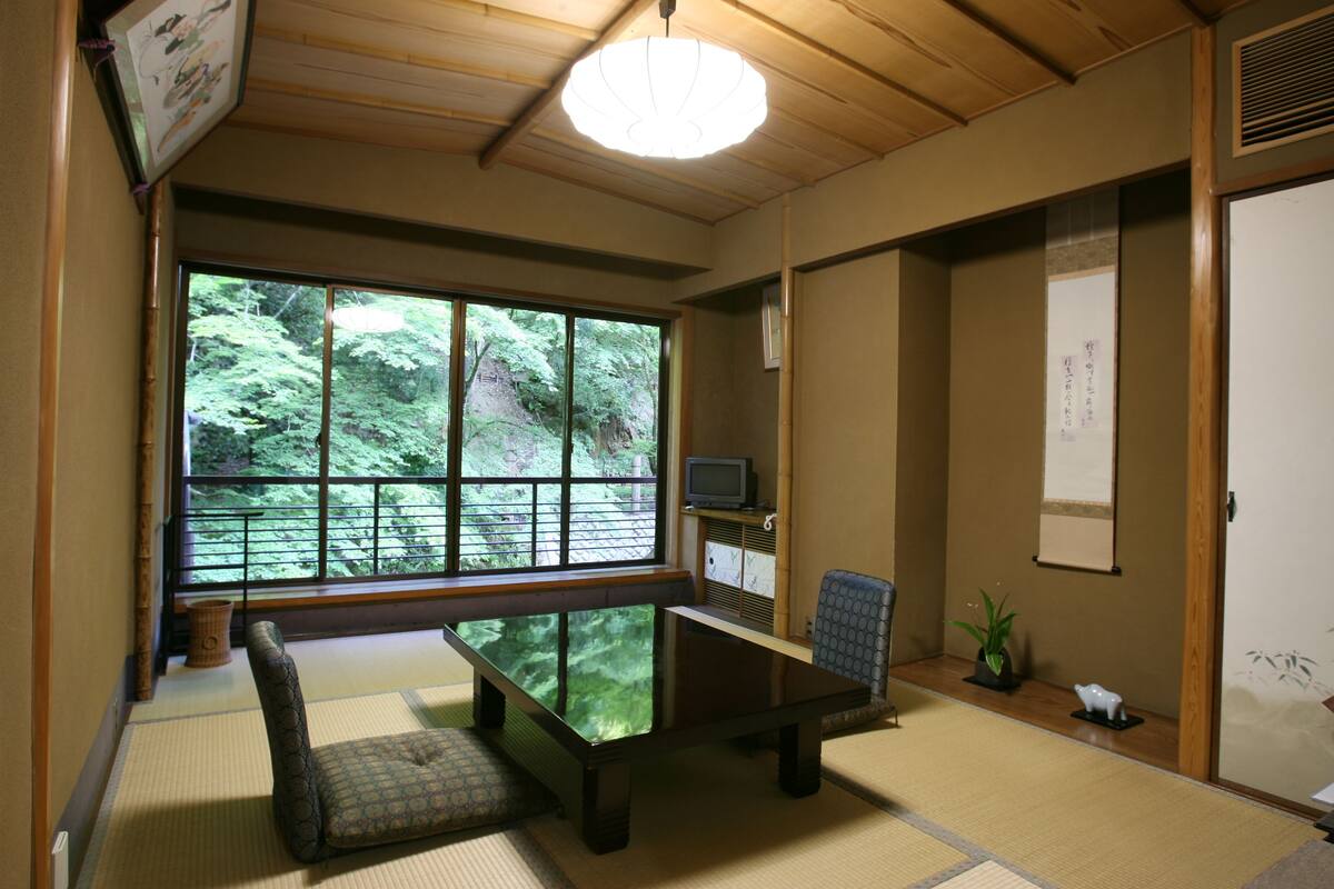 Ein traditioneller japanischer Raum mit Tatami-Böden, einem niedrigen Tisch, einer gepolsterten Sitzbank, einem Fernseher und einem großen Fenster mit Blick auf Grünflächen.