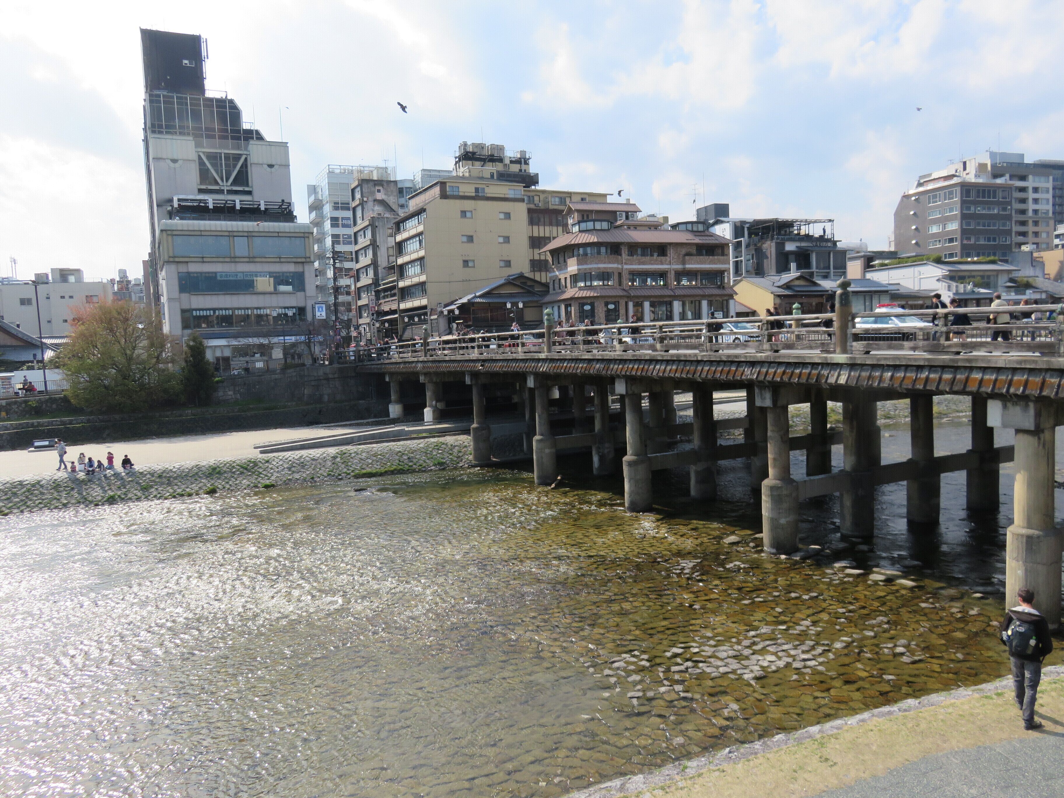 Un paseo a orillas del río con un sendero de piedra, un puente de madera y edificios al fondo.