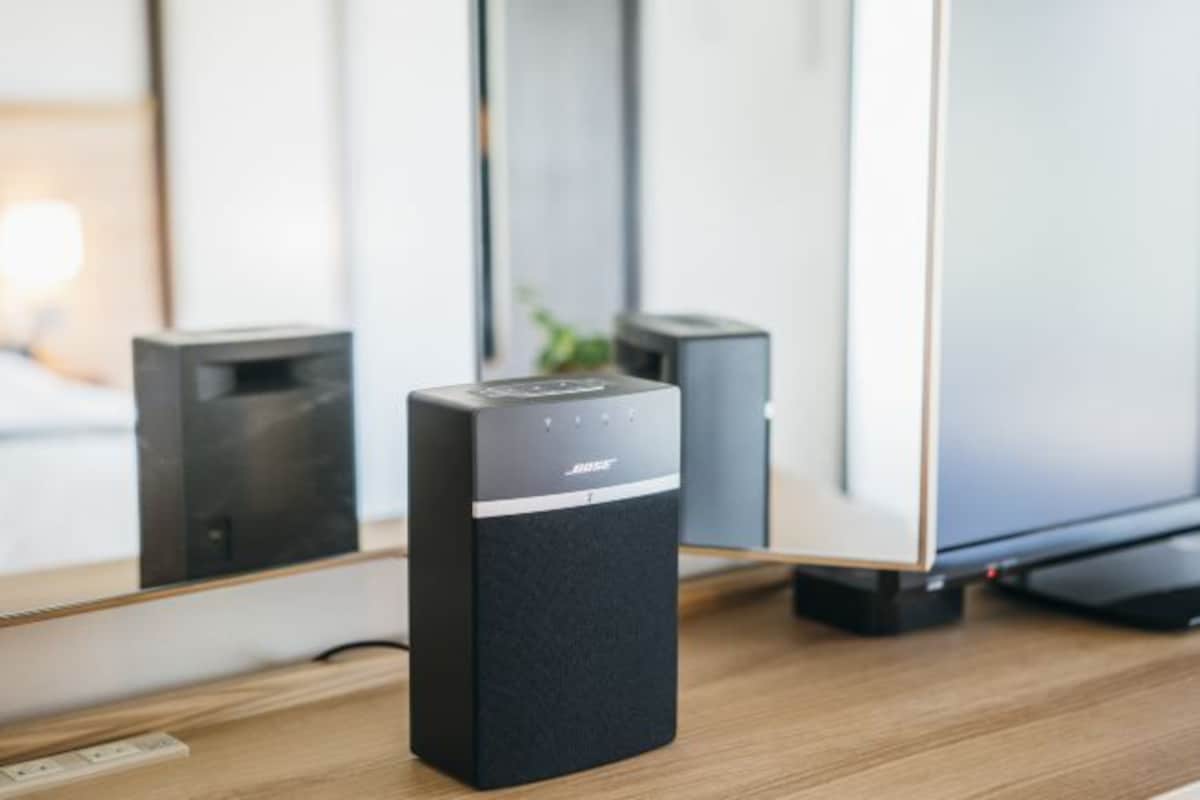 A black Bose speaker on a wooden desk with a mirror reflecting a TV and a plant.