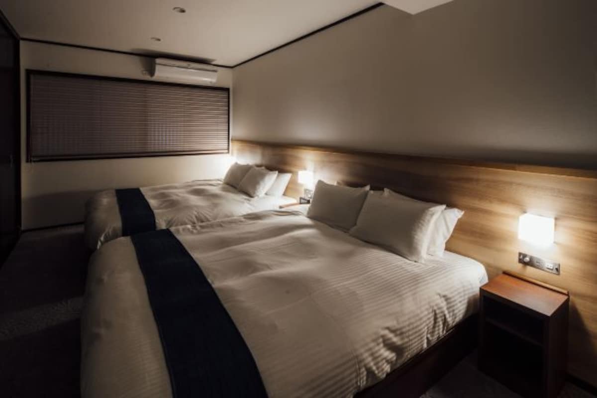 A hotel room with two beds, a wooden headboard, bedside lamps, and a large window with blinds.