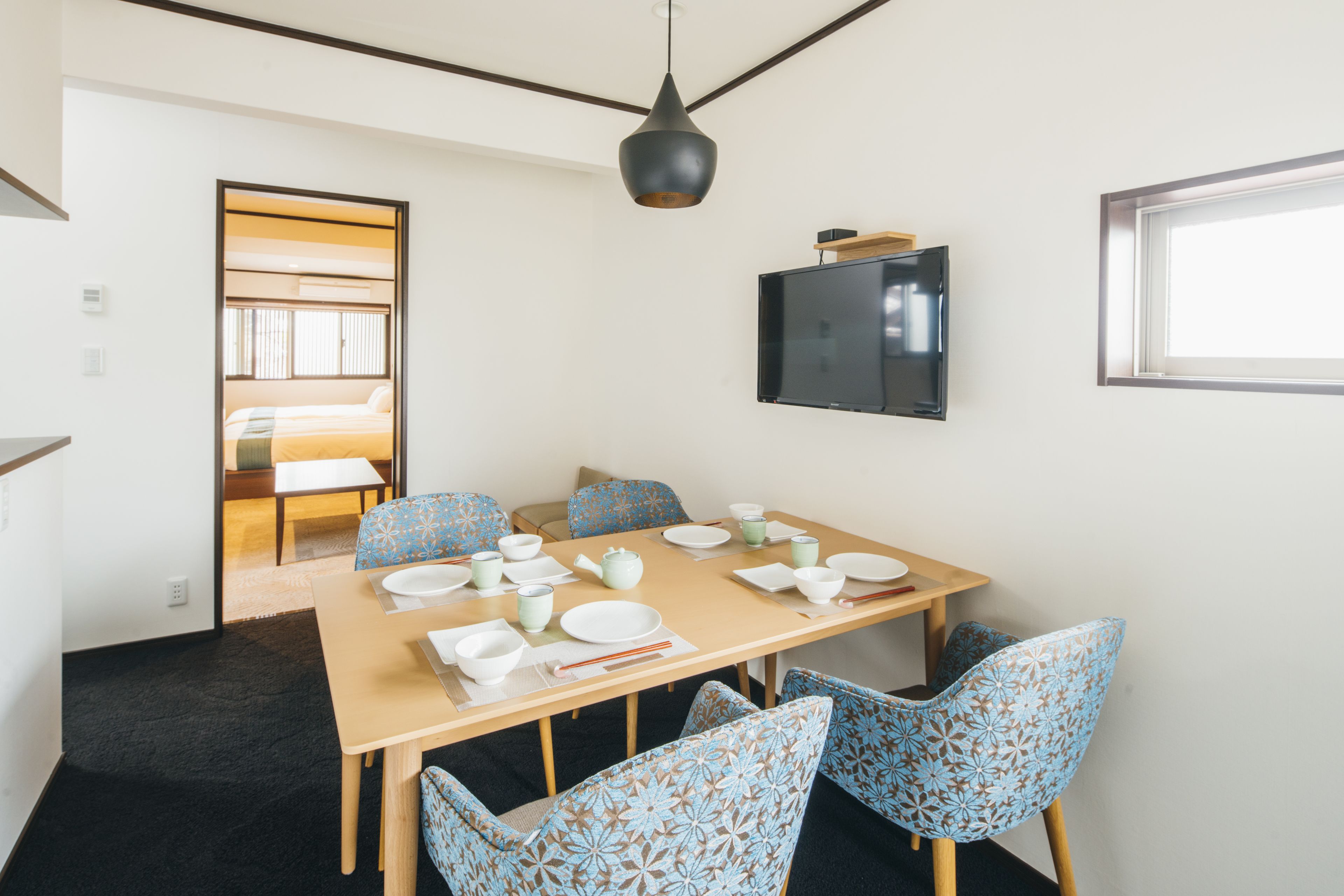 Uma sala de jantar com mesa de madeira posta para quatro pessoas, cercada por cadeiras estampadas, uma televisão na parede e uma janela que oferece luz natural.