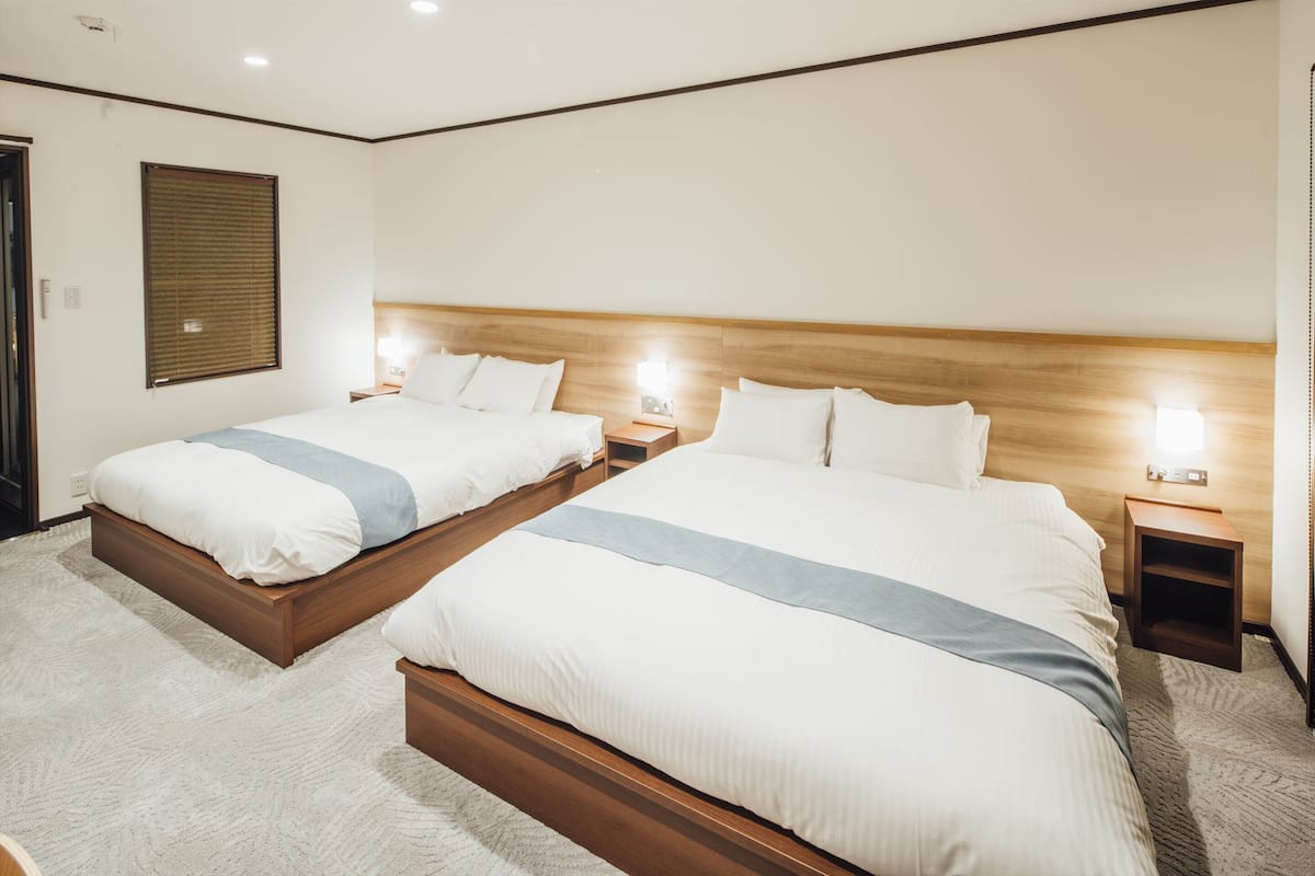 Une chambre d’hôtel avec deux lits, des têtes de lit en bois, des tables de chevet avec des lampes et une grande fenêtre avec des stores.