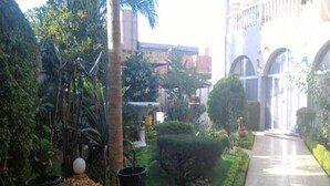 A courtyard with a paved walkway, a small garden area with a tree and a spherical object, and a building with arches in the background. - Residence Diane (Ouagadougou)