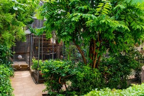 A lush garden with a variety of plants and trees, a tiled pathway, and a fenced area with a birdcage. - Residence Diane (Ouagadougou)