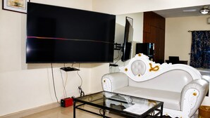A living room with a flat-screen TV mounted on the wall, a glass coffee table, a white sofa, and a wooden cabinet.