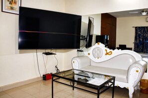 Una sala de estar con un televisor de pantalla plana montado en la pared, una mesa de centro de vidrio, un sofá blanco y un armario de madera.