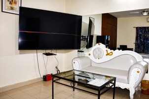 A living room with a flat-screen TV mounted on the wall, a glass coffee table, a white sofa, and a wooden cabinet.