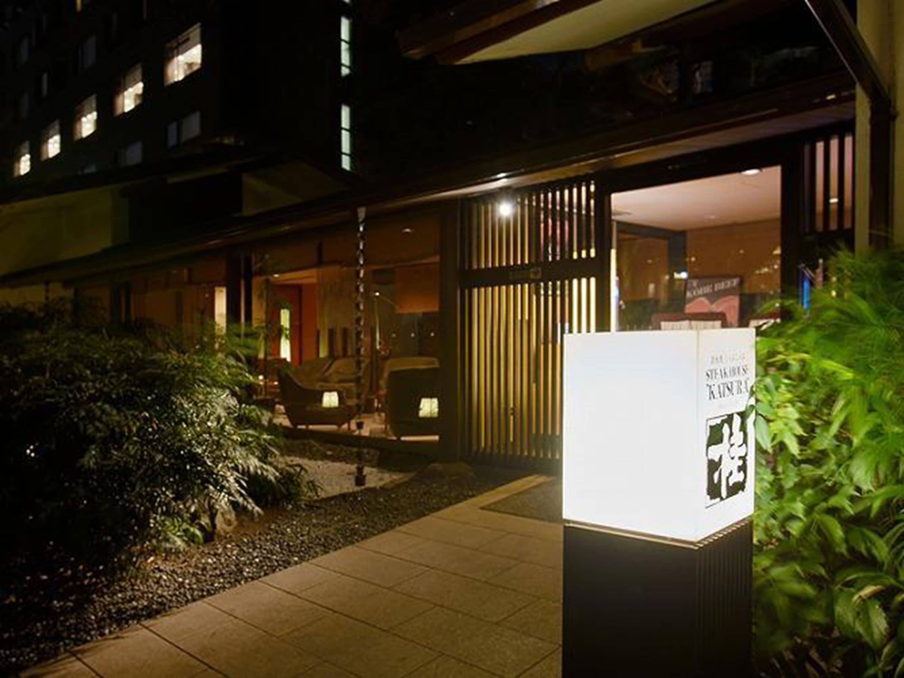 A hotel entrance with a modern design, featuring a pathway leading to the entrance, a seating area with chairs, and a signboard.