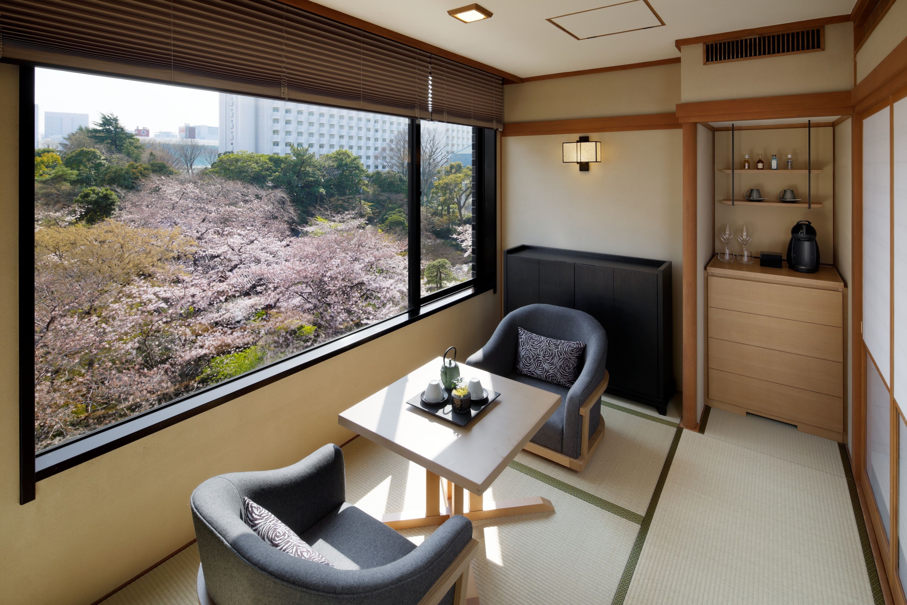 A room with a view of cherry blossoms, a table with a tray, two chairs, a cabinet with bottles, and a kettle.