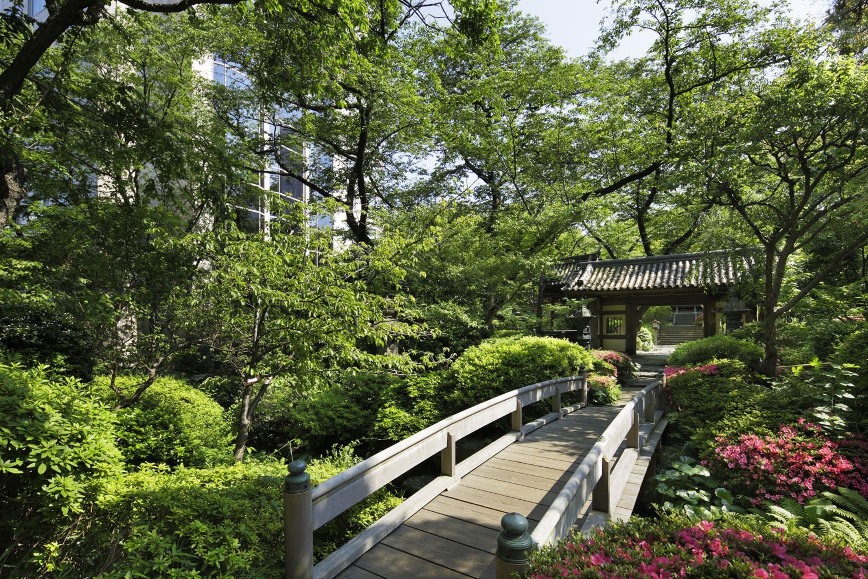 A wooden bridge in a lush garden with dense greenery and a traditional building in the background.
