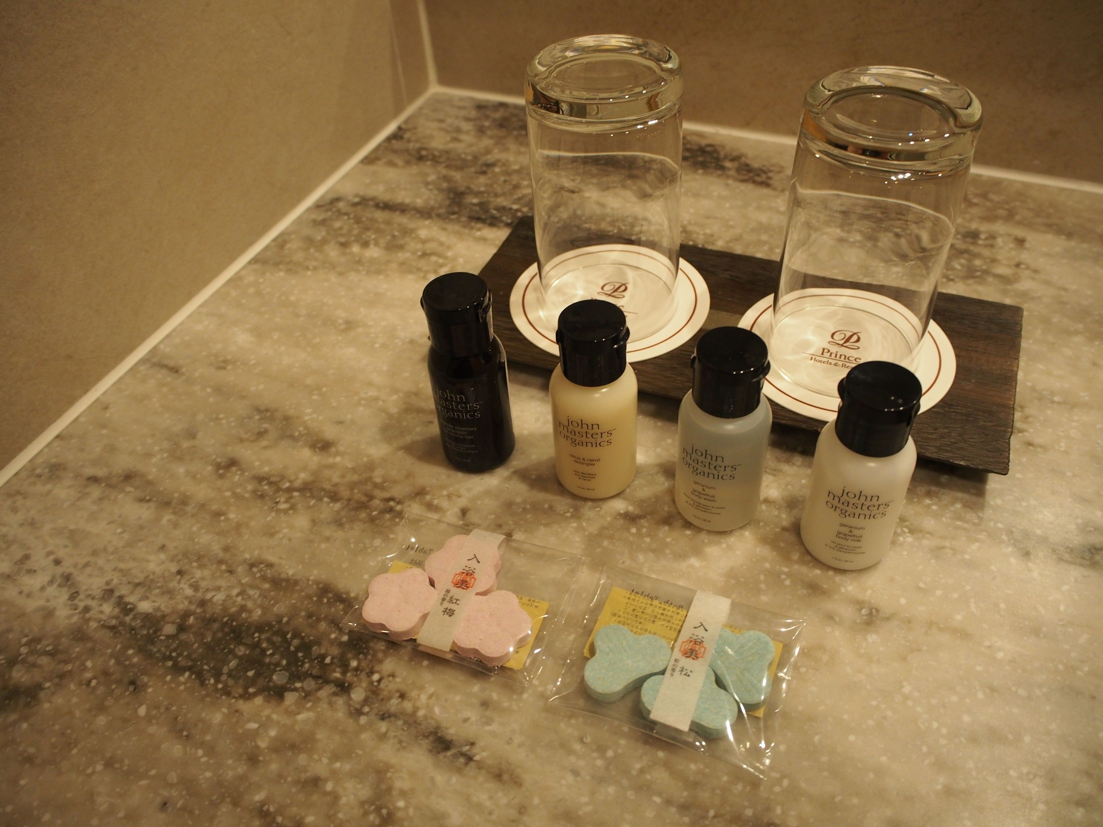 A bathroom counter with toiletries including bottles of John Masters Organics products, a glass jar, and packaged soap bars.