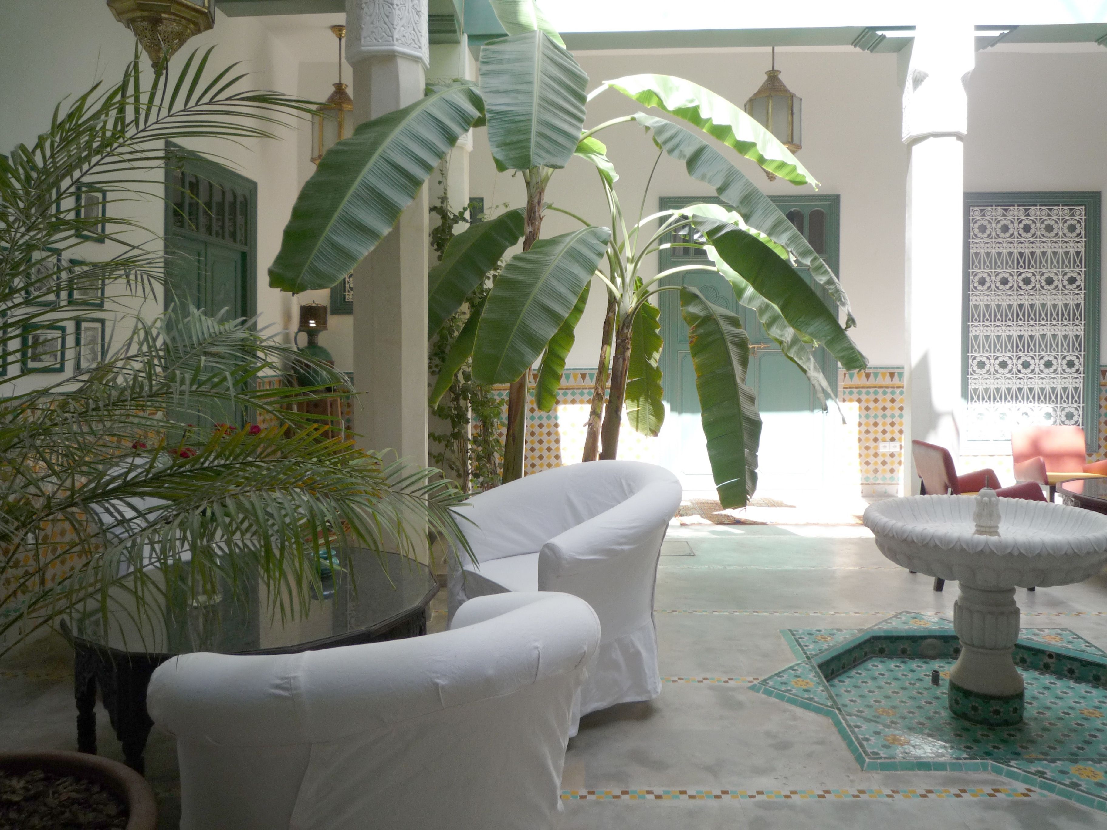 A courtyard with white chairs, a table, and a decorative fountain. There are large green plants and a tiled floor.