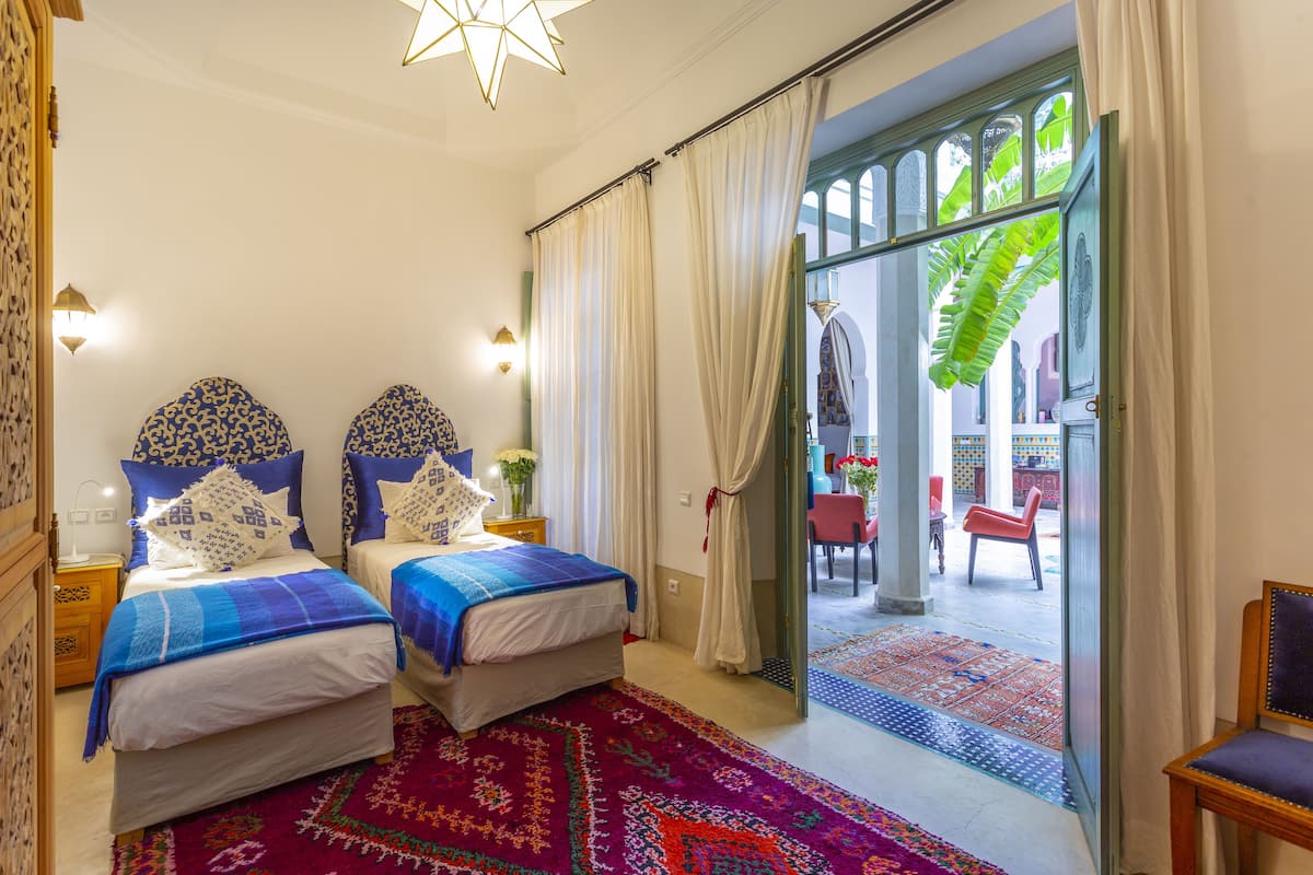 A hotel room with two beds, a large window with curtains, a view of the courtyard, and a wooden chair.