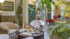 A cozy indoor seating area with a round table set for breakfast, surrounded by comfortable chairs and lush greenery. - Riad Azahar (Marrakech)