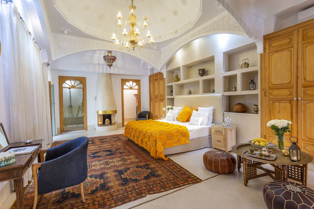 A spacious bedroom with a large bed, a desk, and a chair. There is a chandelier, a wooden cabinet, and a small table with decorative items.