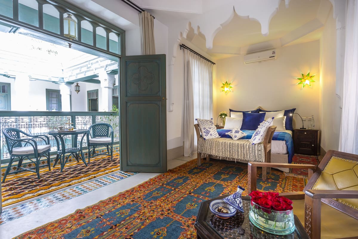 A room with a balcony, a bed with blue bedding, a nightstand, and a decorative vase with red roses.