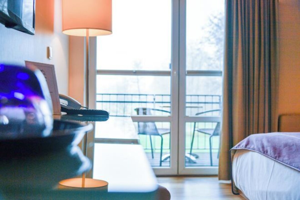 A hotel room with a bed, a desk with a lamp, and a view of an outdoor balcony with chairs.