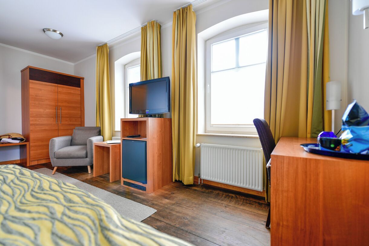 A hotel room with a bed, a desk with a computer, a TV, a wardrobe, and a window with curtains.